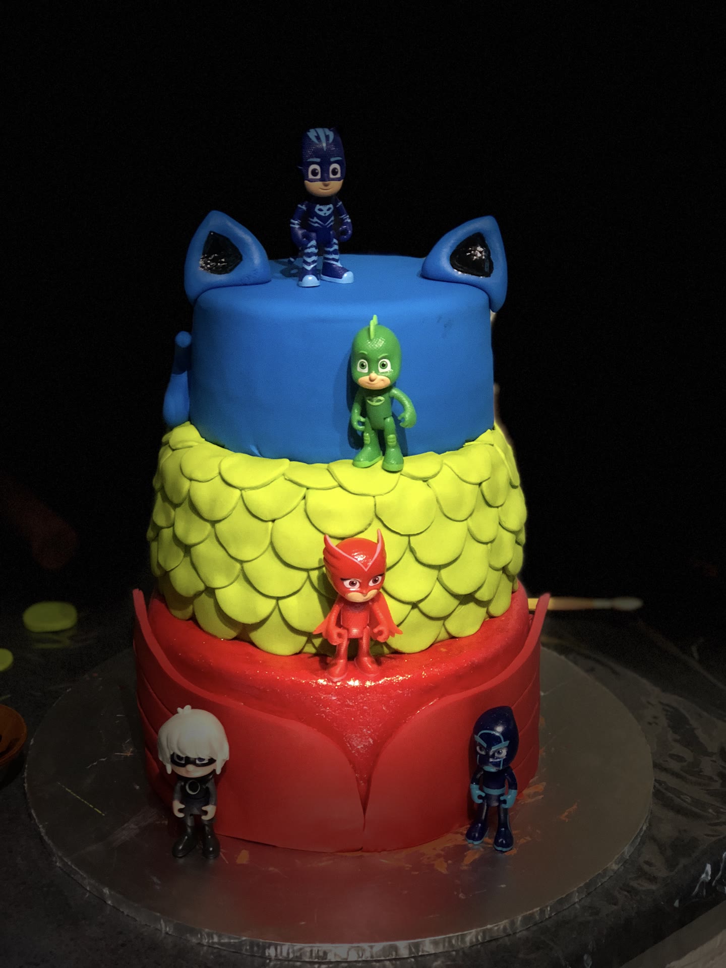 Three tier cake with hand cut green scales and a blue sculpted mask on top.